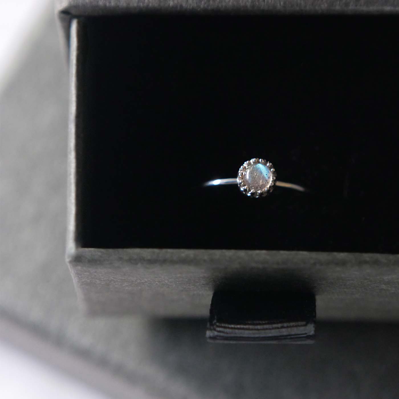 Top front view of a dainty adjustable sterling silver ring with a focal natural high quality aaa labradorite gemstone cabochon set in a crown bezel setting. The ring is tucked into the black velvet insert of a gray textured colored jewelry box.