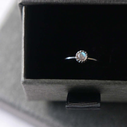 Top front view of a dainty adjustable sterling silver ring with a focal natural high quality aaa labradorite gemstone cabochon set in a crown bezel setting. The ring is tucked into the black velvet insert of a gray textured colored jewelry box.