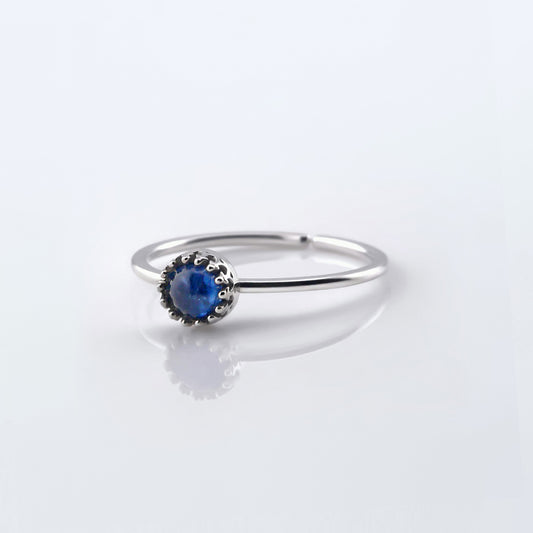 Front view of a dainty adjustable sterling silver ring with a focal blue kyanite solitaire gemstone cabochon set in a crown bezel setting. The ring is laying flat on a plain white reflective surface.