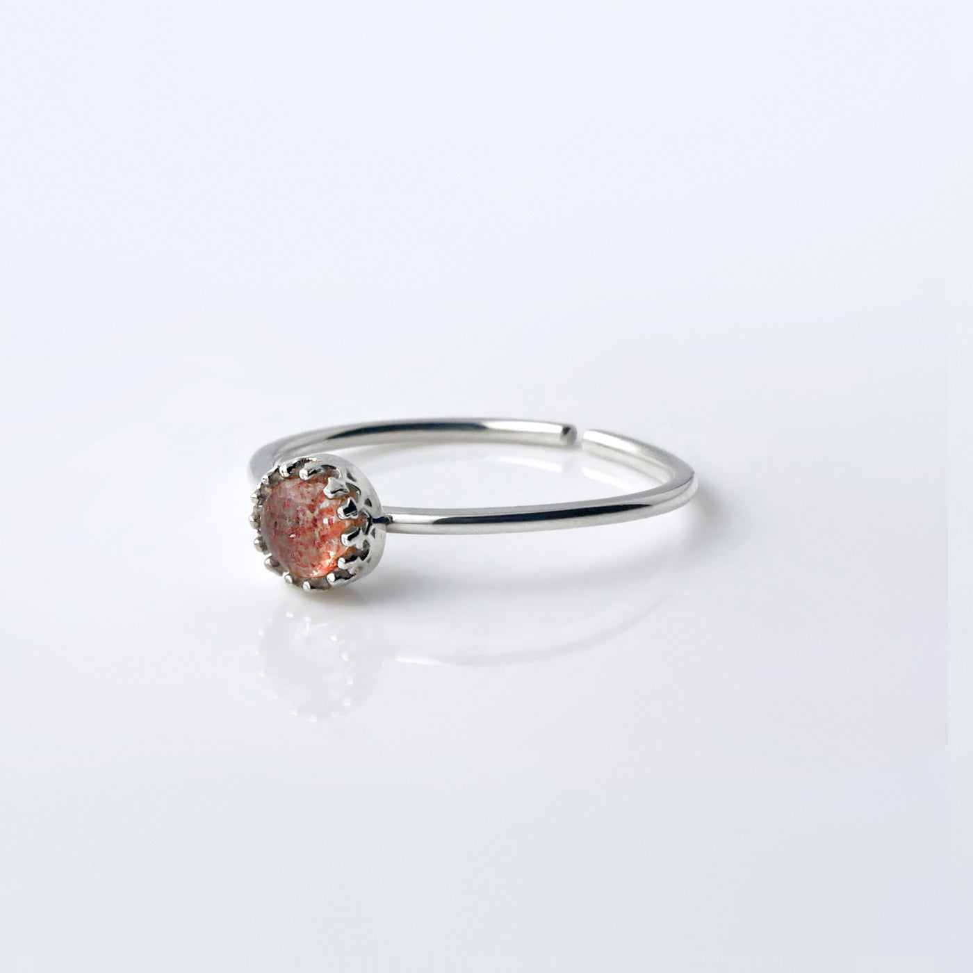Front view of a dainty adjustable sterling silver ring with a focal strawberry quartz solitaire gemstone cabochon set in a crown bezel setting. The ring is laying flat on a plain white reflective surface.