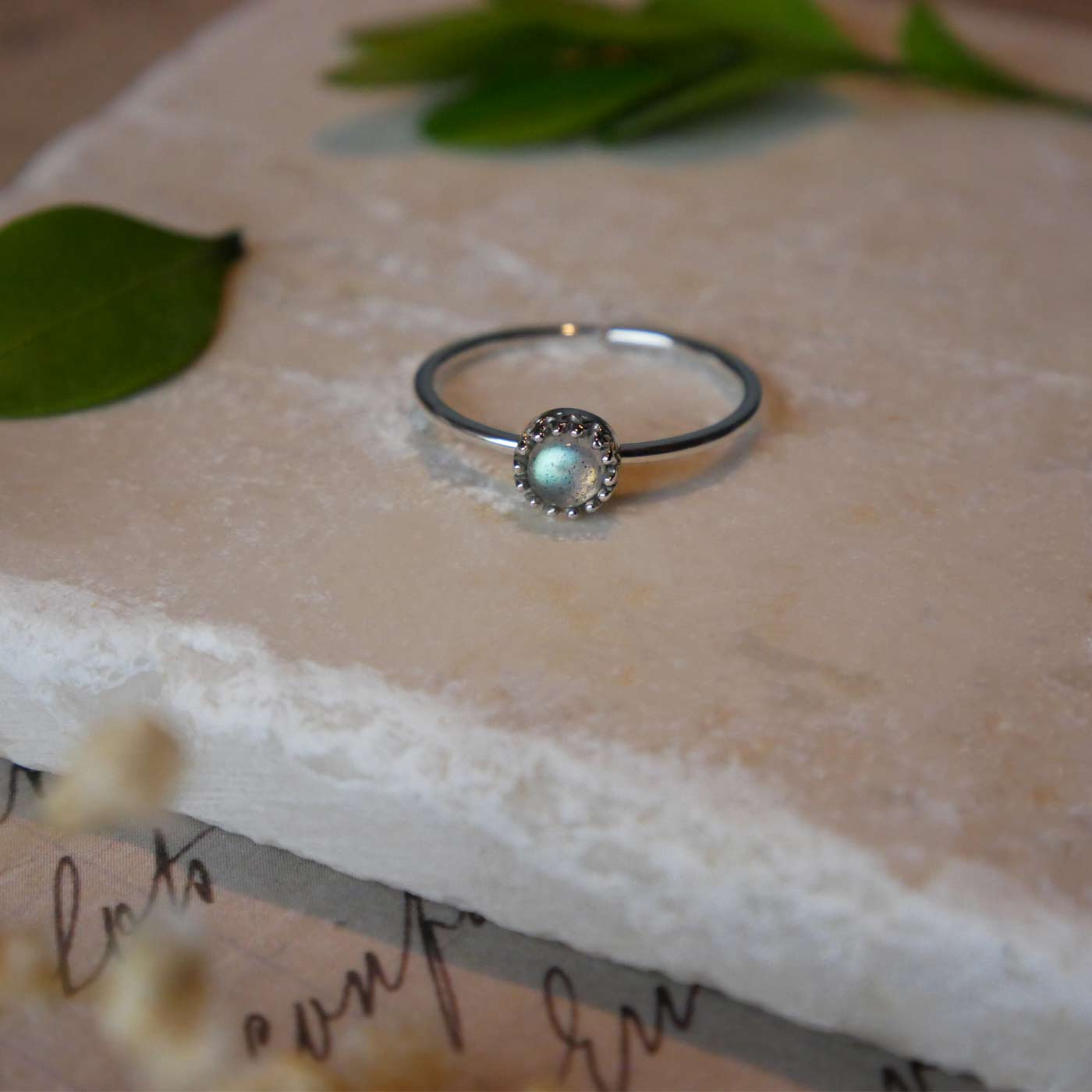 Front view of a dainty adjustable sterling silver ring with a focal natural high quality aaa labradorite gemstone cabochon set in a crown bezel setting laying on a stone surface over textured vintage beige pink paper with cursive writing on it, also stylized with green leaves and blurred beige flowers in the foreground.