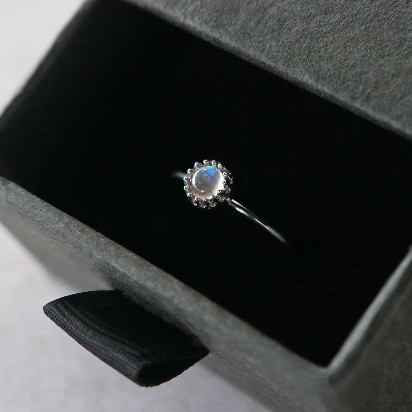 Top 3/4 view of a dainty adjustable sterling silver ring with a focal natural high quality aaa labradorite gemstone cabochon set in a crown bezel setting. The ring is tucked into the black velvet insert of a gray textured colored jewelry box.