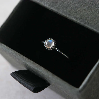 Top 3/4 view of a dainty adjustable sterling silver ring with a focal natural high quality aaa labradorite gemstone cabochon set in a crown bezel setting. The ring is tucked into the black velvet insert of a gray textured colored jewelry box.
