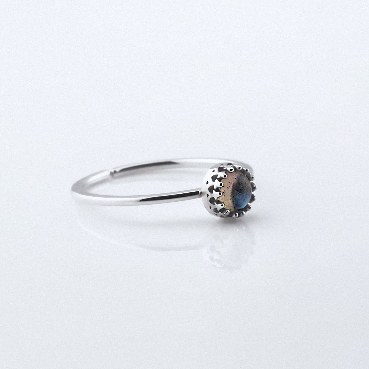 3/4 view of a dainty adjustable sterling silver ring with a focal high quality natural AAA labradorite gemstone cabochon set in a crown bezel setting. The ring is laying flat on a plain white reflective surface.