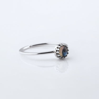 3/4 view of a dainty adjustable sterling silver ring with a focal high quality natural AAA labradorite gemstone cabochon set in a crown bezel setting. The ring is laying flat on a plain white reflective surface.