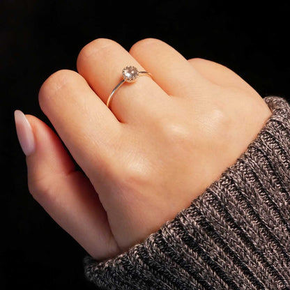 Hand wearing a dainty adjustable sterling silver ring with a focal natural high quality aaa labradorite gemstone cabochon set in a crown bezel setting. The person has a gray textured sleeve on with a dark black background.