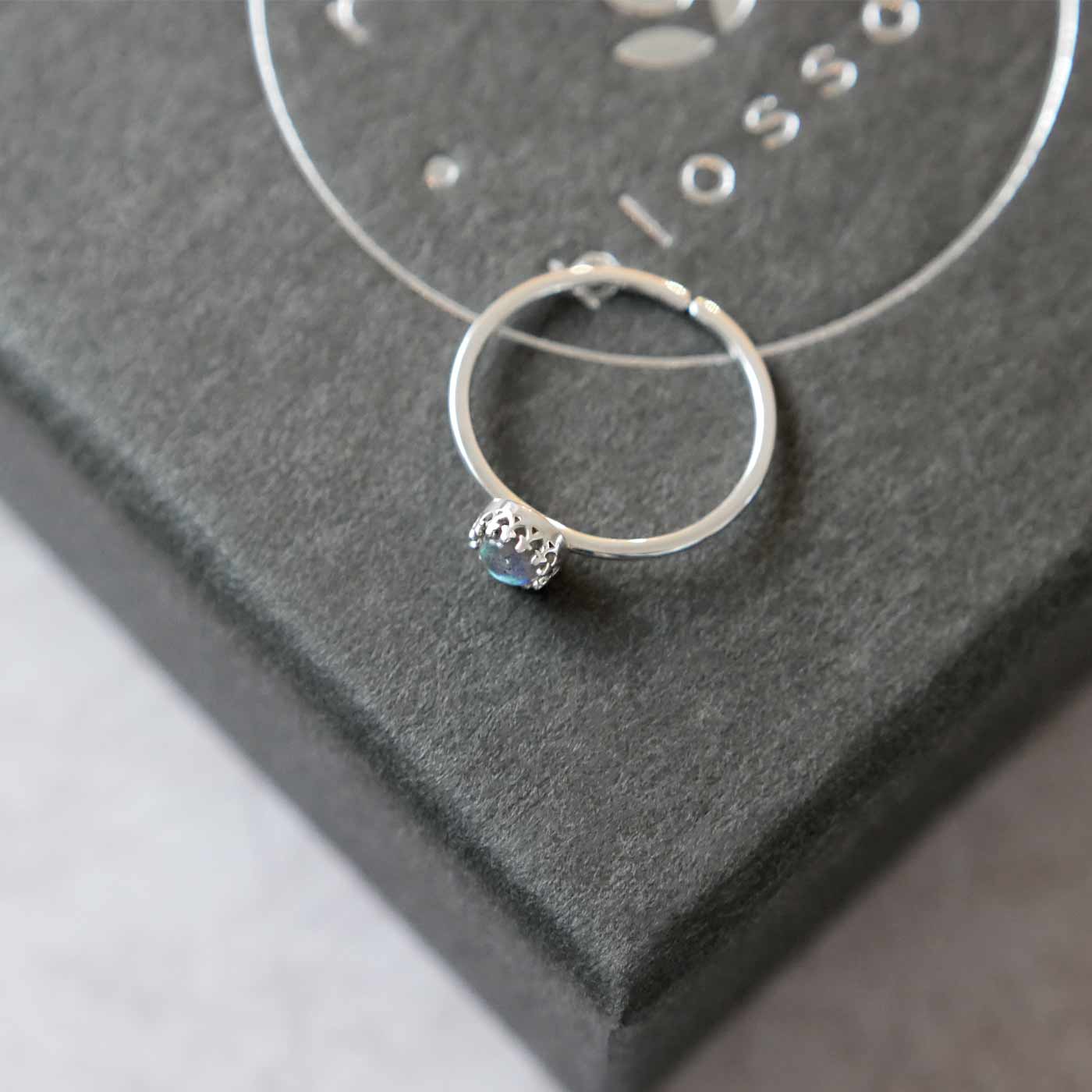 Top side view of a dainty adjustable sterling silver ring with a focal natural aaa labradorite gemstone cabochon set in a crown bezel setting. The ring is laying flat on a gray textured colored jewelry box with silver reverie blossom branded text.