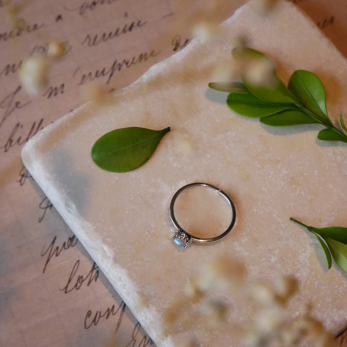 Top view of a dainty adjustable sterling silver ring with a focal natural high quality aaa labradorite gemstone cabochon set in a crown bezel setting laying on a stone surface over textured vintage beige pink paper with cursive writing on it, also stylized with green leaves and blurred beige flowers in the foreground.