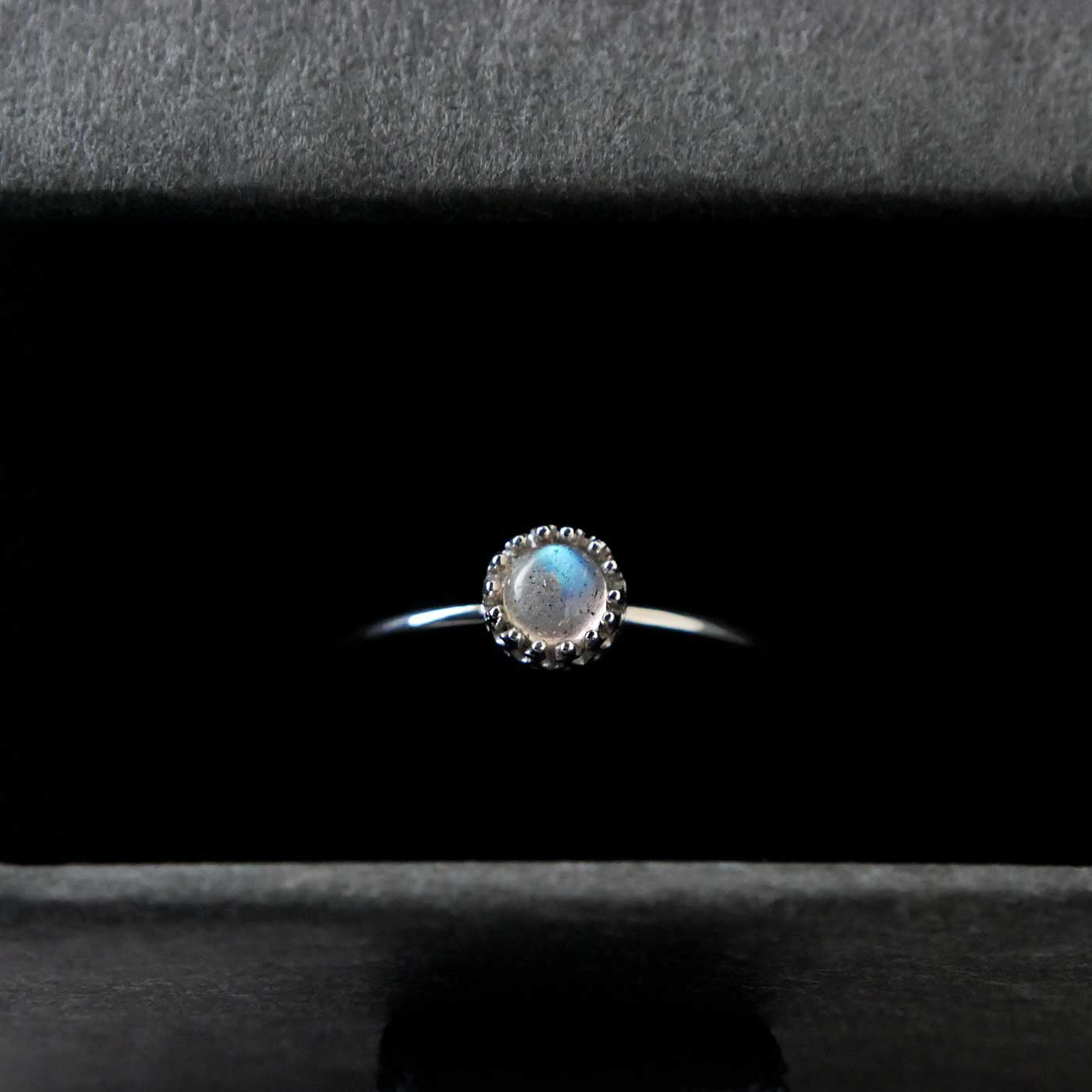 Top view of a dainty adjustable sterling silver ring with a focal natural high quality aaa labradorite gemstone cabochon set in a crown bezel setting. The ring is tucked into the black velvet insert of a gray textured colored jewelry box.