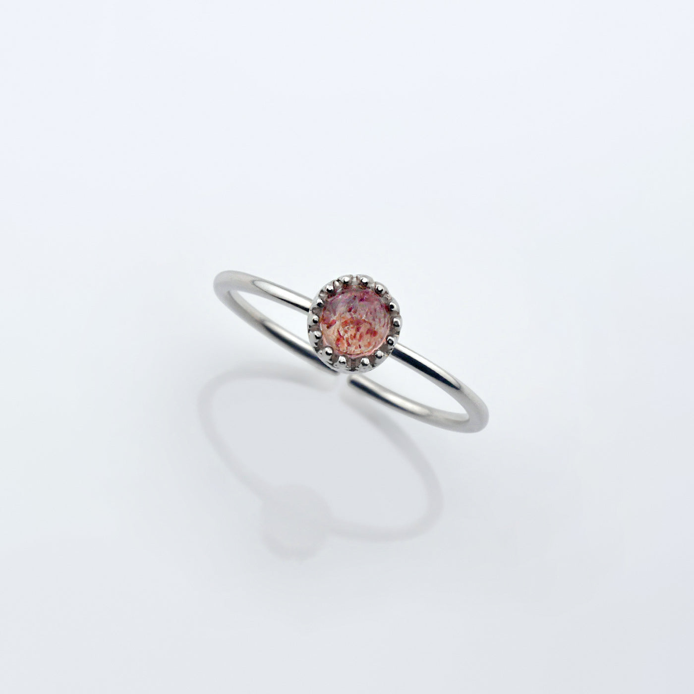 Top view of a dainty adjustable sterling silver ring with a focal strawberry quartz solitaire gemstone cabochon set in a crown bezel setting. The ring is elevated on a plain white reflective surface.