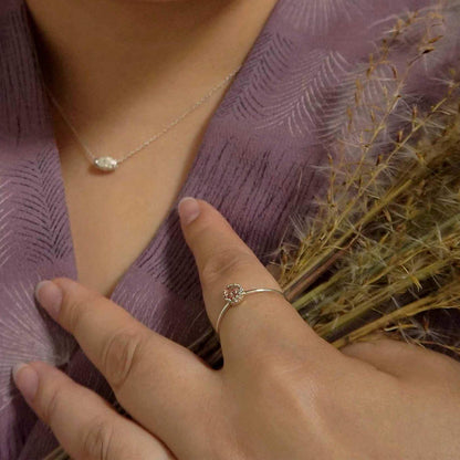 Close-up of a hand wearing a dainty adjustable sterling silver ring with a focal strawberry quartz solitaire gemstone cabochon set in a crown bezel setting. The person is holding natural plants in one hand, also wearing a dainty sterling silver barrel necklace, and a purple Japanese textured garment in the background.