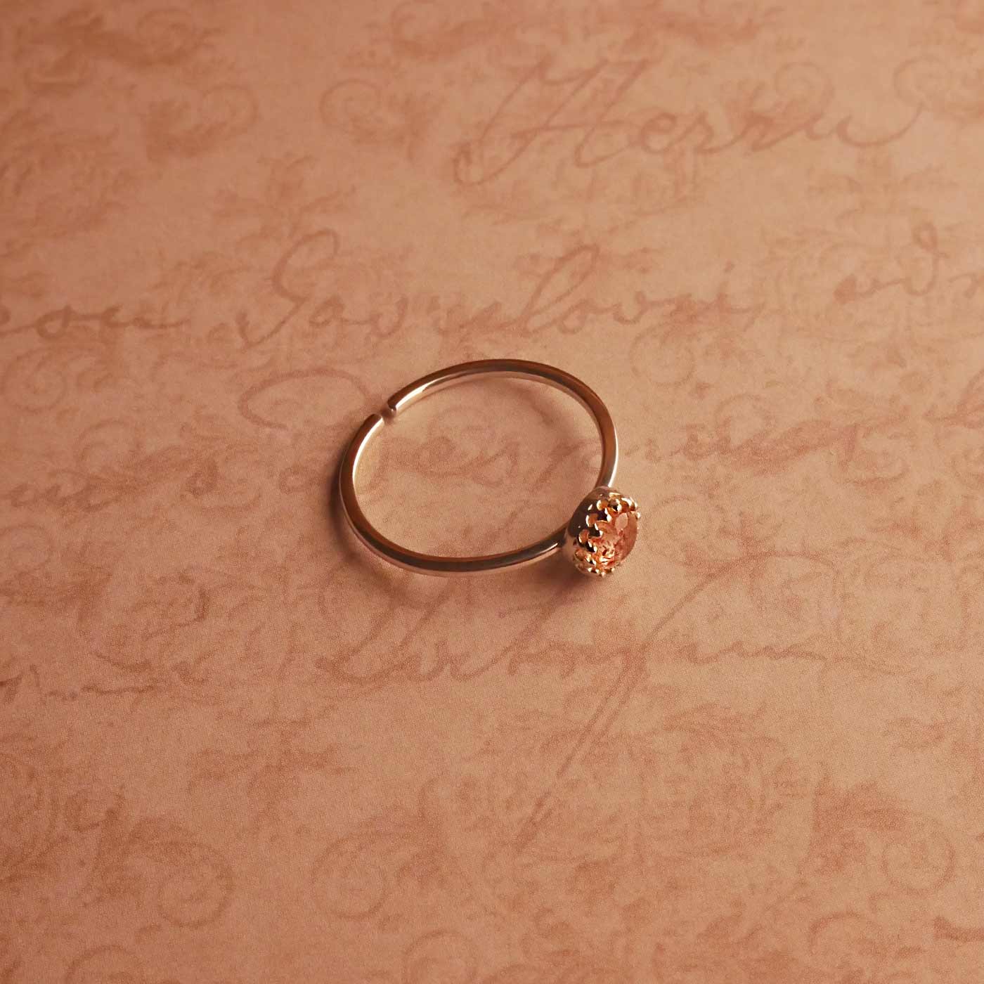 Top angle side view of a dainty adjustable sterling silver ring with a focal strawberry quartz solitaire gemstone cabochon, set in a crown bezel setting. The ring is laying flat over a textured beige pink vintage paper background with cursive writing on it.