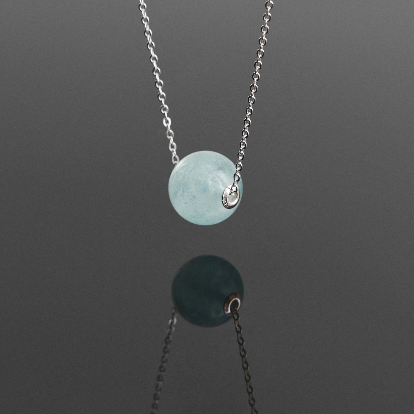 3/4 front view of a dainty round aquaramine ball sphere pendant necklace with sterling silver 925 rivets on a sterling silver chain hanging, with a plain reflective dark gray background.