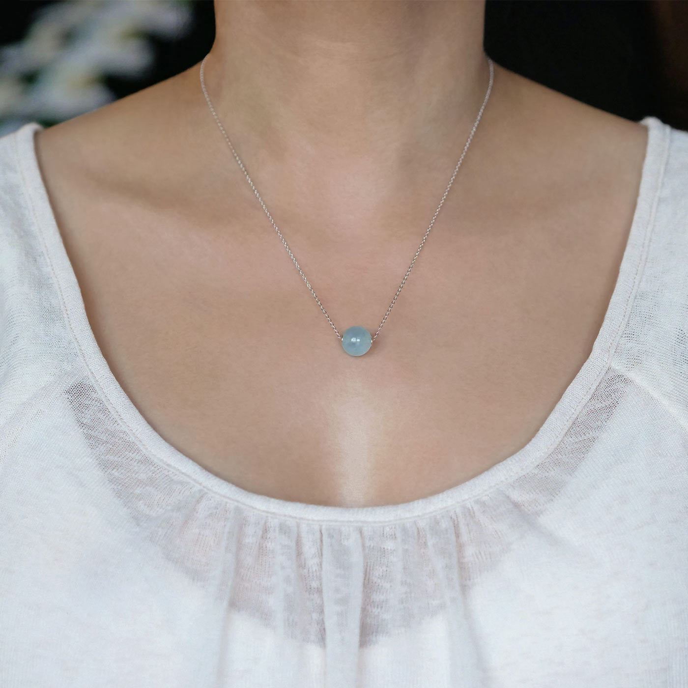 Front view of a dainty, round aquamarine ball sphere with sterling silver rivets pendant chain necklace worn by a person with a beige top, against a blurred dark background with white flowers.