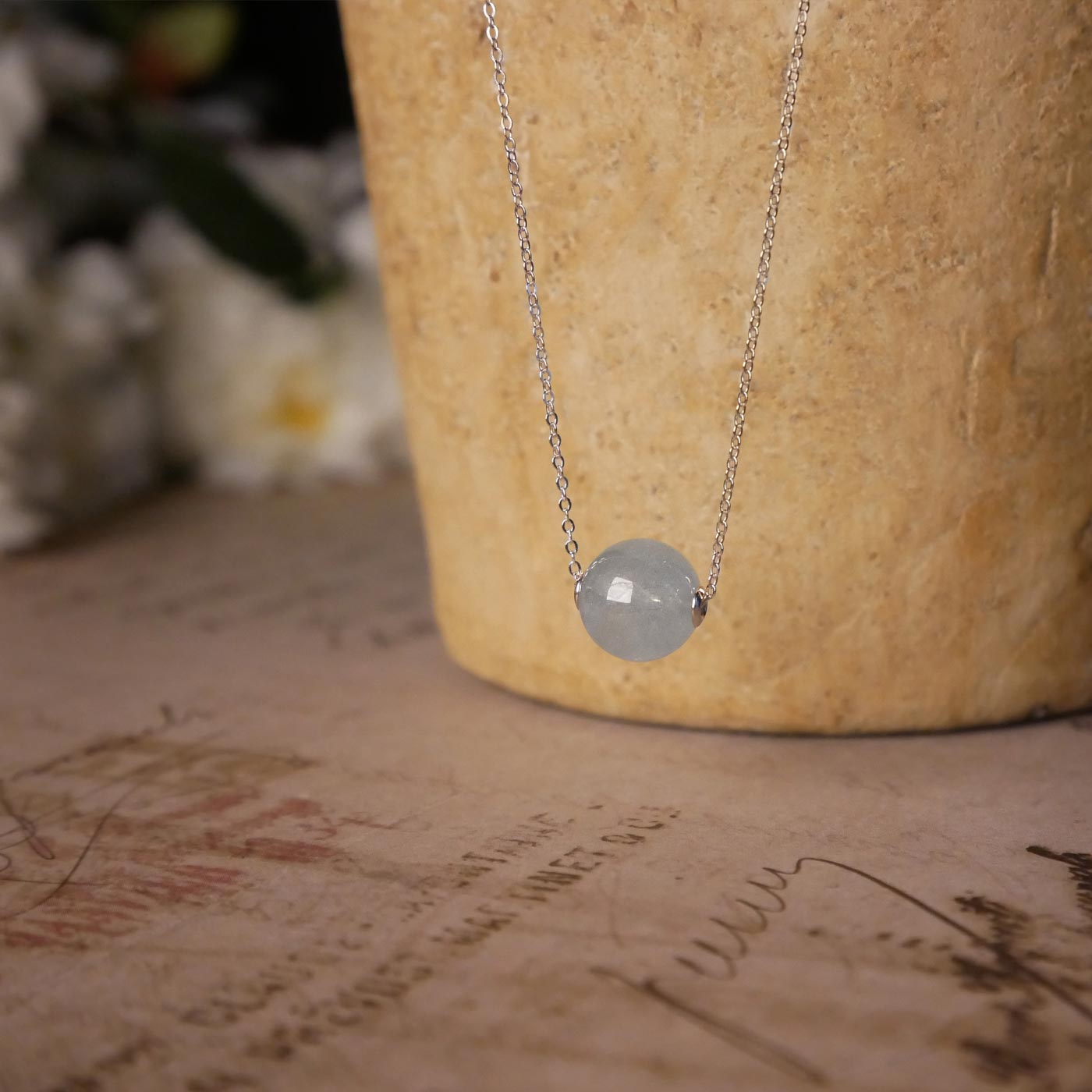 Front view of a dainty, round aquamarine ball sphere with sterling silver rivets pendant chain necklace. It is stylized by hanging from a textured light brown pot, over vintage paper with old writing style on it, with white flowers blurred in background.
