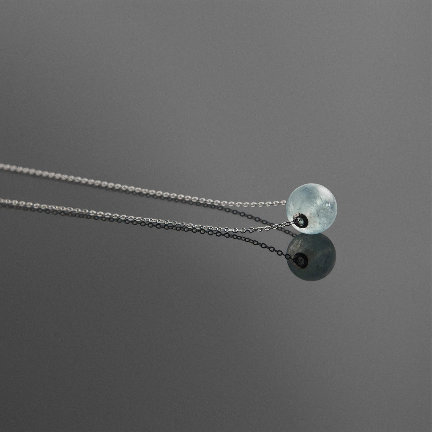 Side view of a dainty round aquaramine ball sphere pendant necklace with sterling silver 925 rivets on a sterling silver chain, laying flat on a plain reflective dark gray background.