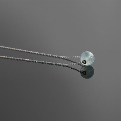 Side view of a dainty round aquaramine ball sphere pendant necklace with sterling silver 925 rivets on a sterling silver chain, laying flat on a plain reflective dark gray background.