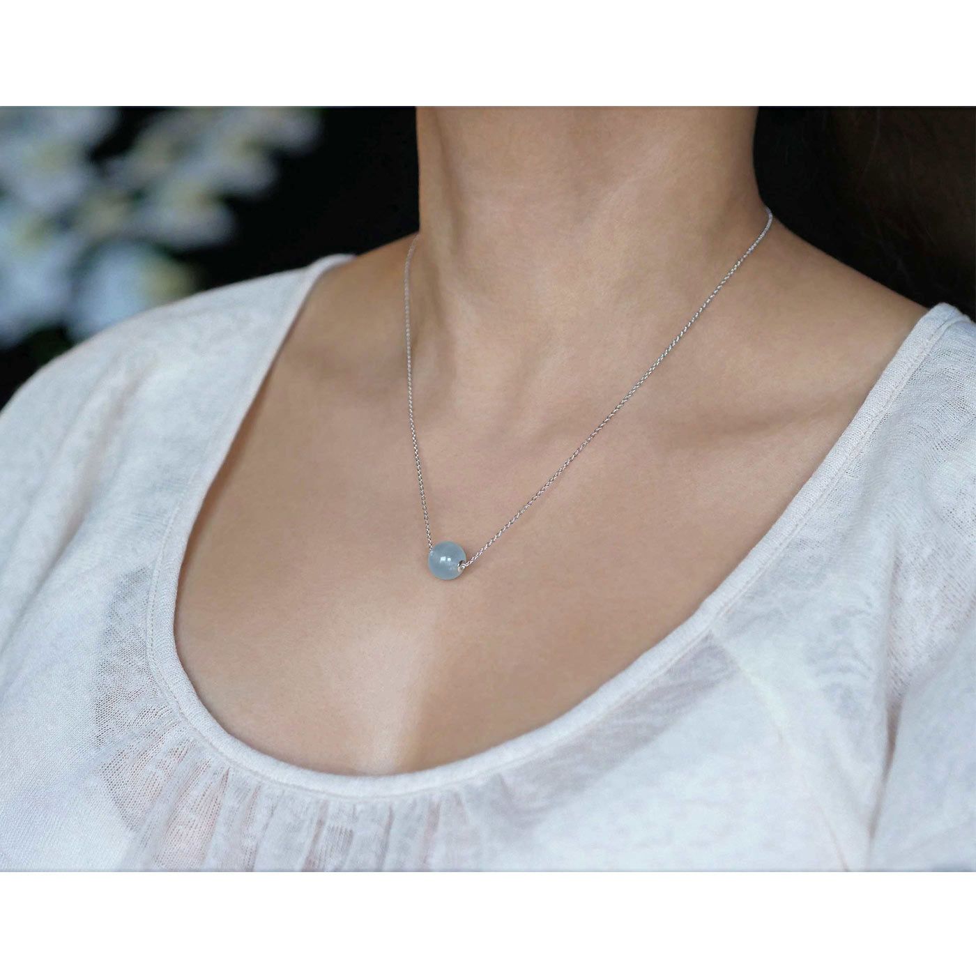3/4 front view of a dainty, round aquamarine ball sphere with sterling silver rivets pendant chain necklace worn by a person with a beige top, against a blurred dark background with white flowers.
