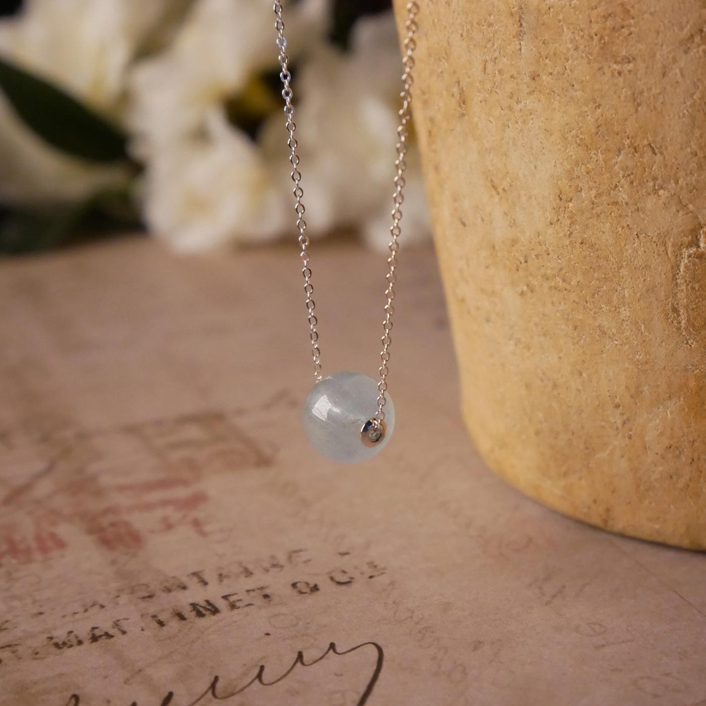 3/4 side top view of a dainty, round aquamarine ball sphere with sterling silver rivets pendant chain necklace. It is stylized by hanging from a textured light brown pot, over vintage paper with old writing style on it, with white flowers blurred in background.