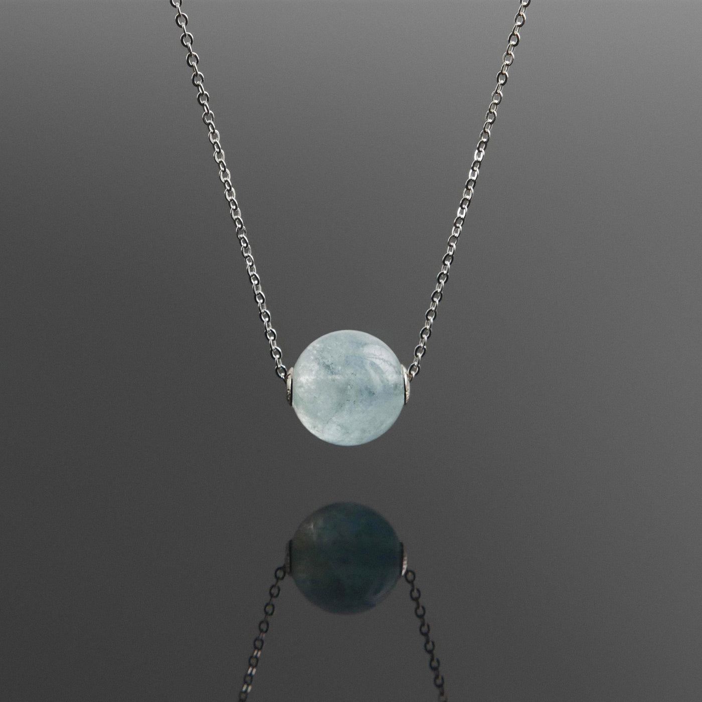 Front view of a dainty round aquaramine ball sphere pendant necklace with sterling silver 925 rivets on a sterling silver chain hanging, with a plain reflective dark gray background.
