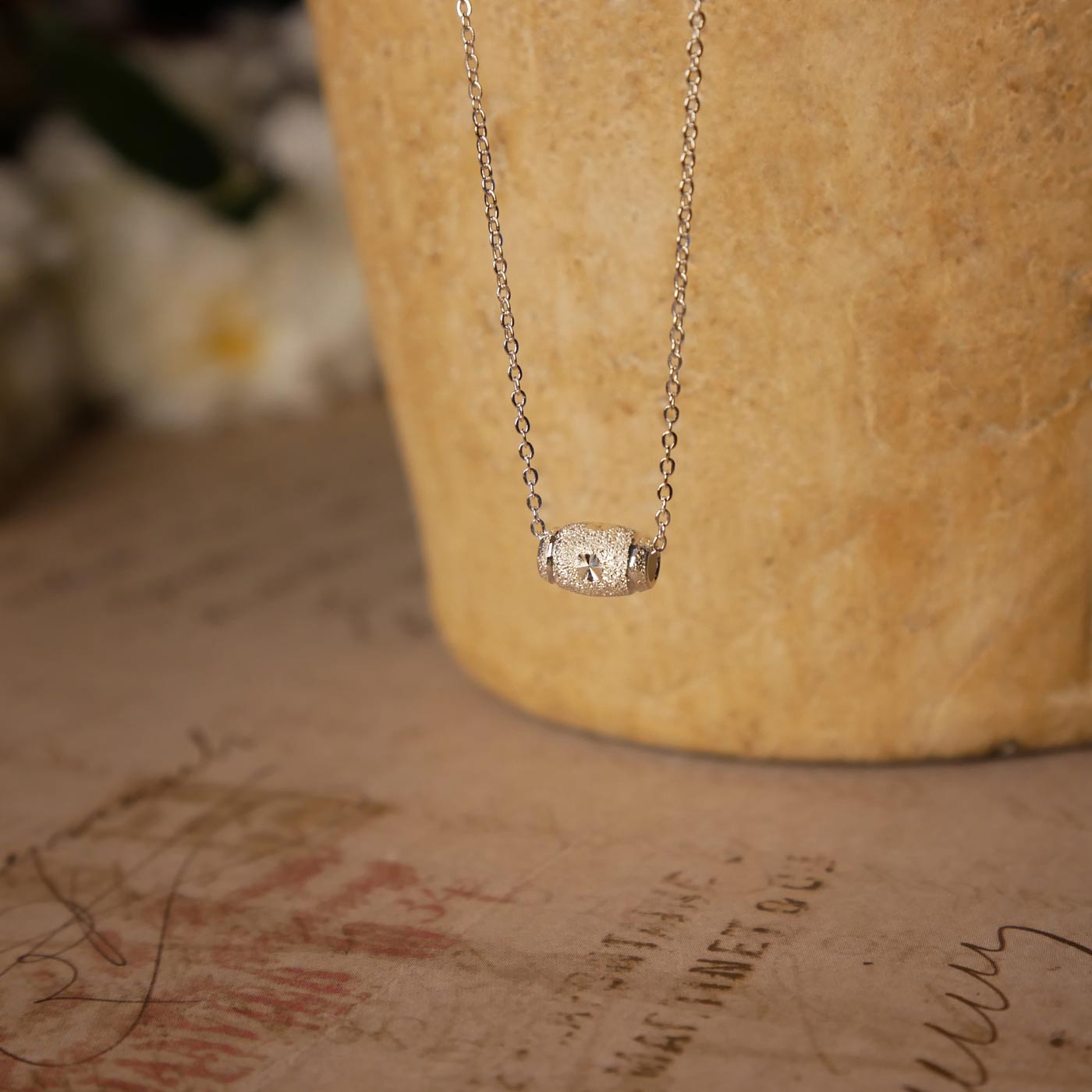 Front view of dainty, sparkly textured sterling silver barrel pendant necklace. It is stylized by hanging from a textured light brown pot, over vintage paper, with old writing style on it and white flowers blurred in the background.
