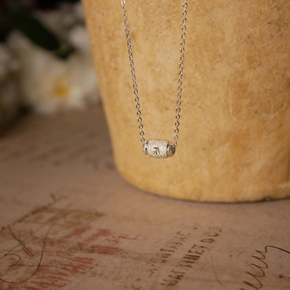 Front view of dainty, sparkly textured sterling silver barrel pendant necklace. It is stylized by hanging from a textured light brown pot, over vintage paper, with old writing style on it and white flowers blurred in the background.