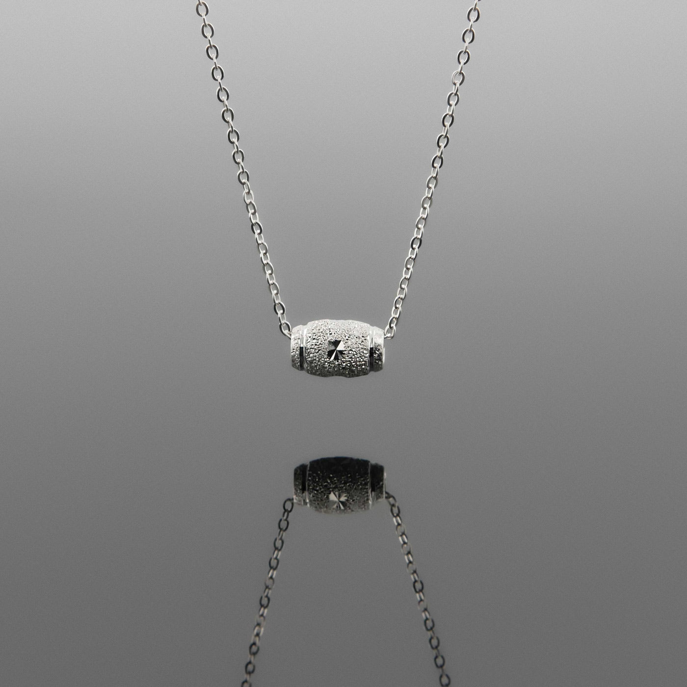Front view of a dainty sparkle textured barrel sterling silver pendant necklace hanging, with a plain reflective gray background.