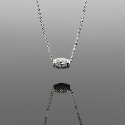 Front view of a dainty sparkle textured barrel sterling silver pendant necklace hanging, with a plain reflective gray background.