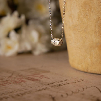 Side and 3/4 view of dainty, sparkly textured sterling silver barrel pendant necklace. It is stylized by hanging from a textured light brown pot, over vintage paper, with old writing style on it and white flowers blurred in the background.