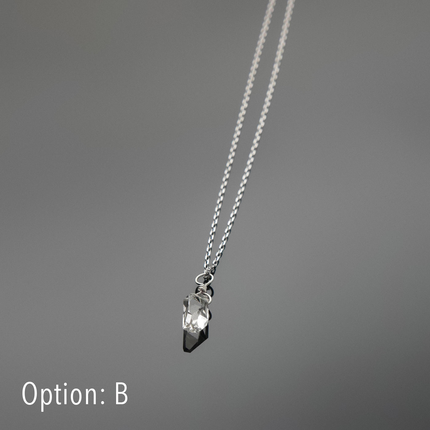 Bottom front view of a dainty, high quality AAA clear raw Herkimer Diamond Quartz wire wrapped sterling silver pendant necklace, laying flat on a plain reflective gray background.