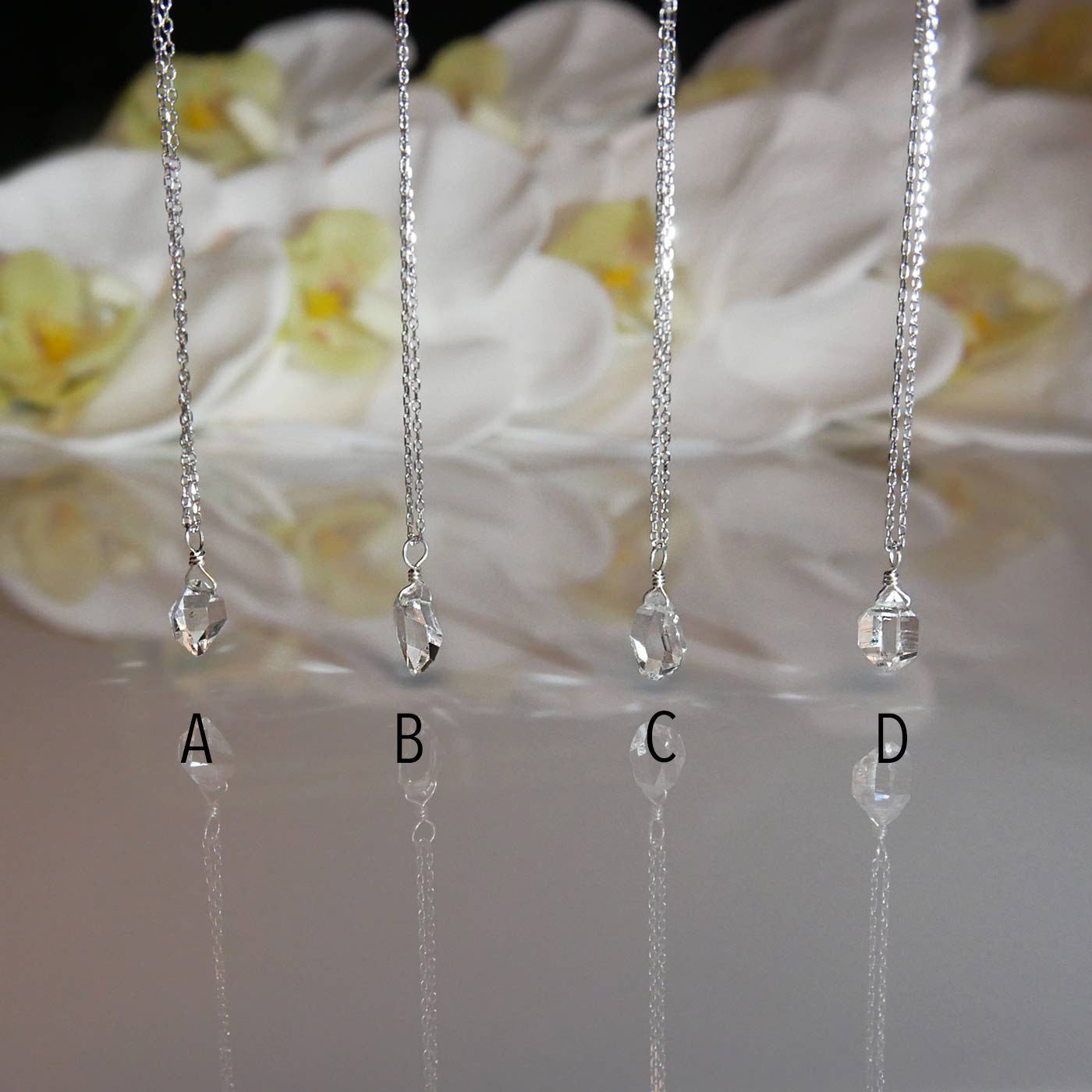Four high quality sparkly AAA dainty Herkimer Diamond Quartz wire wrapped pendant chain necklaces in sterling silver hanging from above. They are stylized hanging over a light reflective surface, with white orchid flowers blurred in the background.