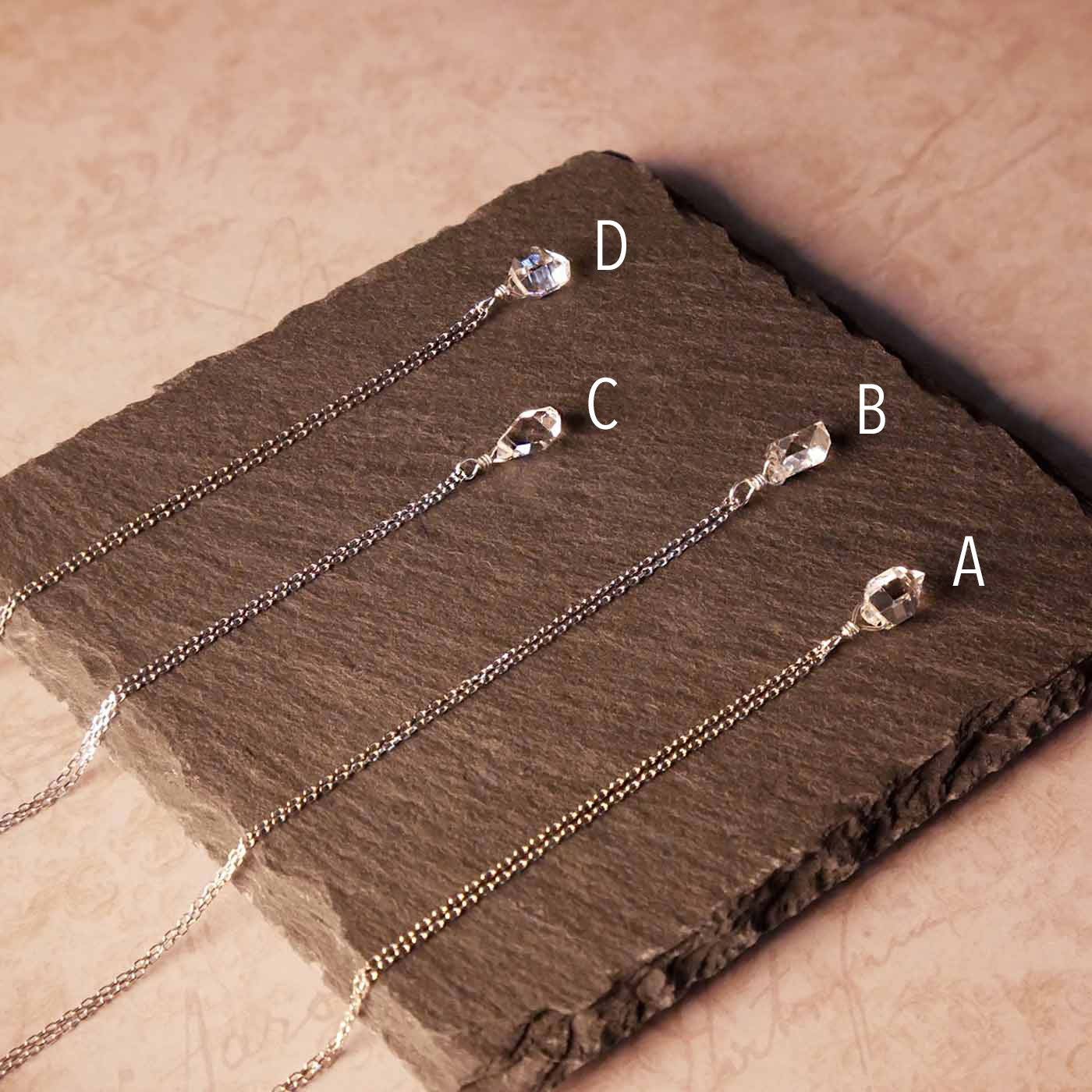 Top back angle view of 4 high quality AAA dainty Herkimer Diamond Quartz wire wrapped pendant chain necklaces in sterling silver. They are stylized laying flat on a dark gray slate surface with vintage pink beige paper underneath.