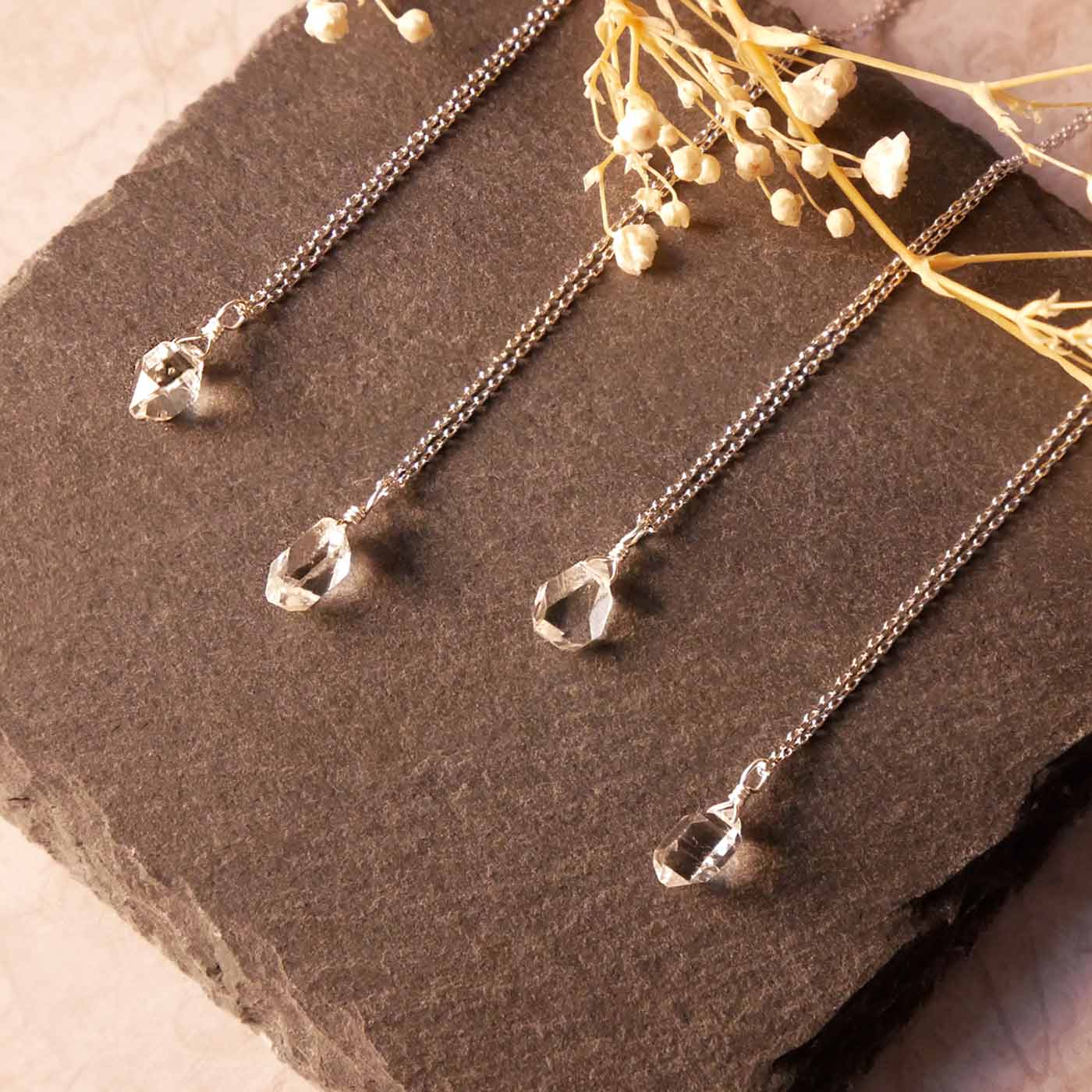 Top angle view of 4 high quality AAA dainty Herkimer Diamond Quartz wire wrapped pendant chain necklaces in sterling silver. They are stylized laying flat on a dark gray slate surface with dried baby's breath flowers in the foreground.