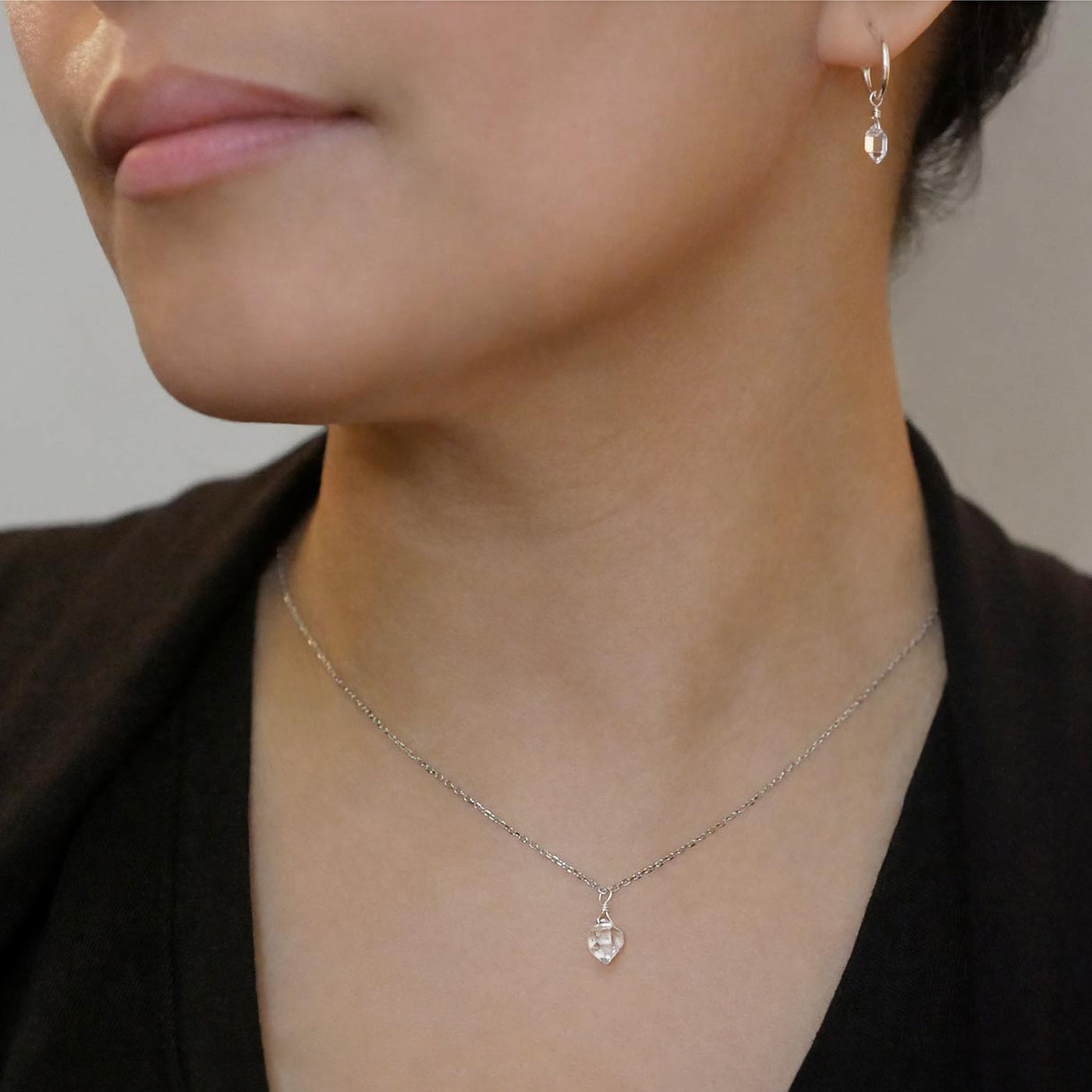 Close-up of a person wearing a dainty high quality AAA Herkimer Diamond Quartz wire wrapped pendant chain necklace and matching Herkimer earring hoops in sterling silver. The person has on a black top and sweater.