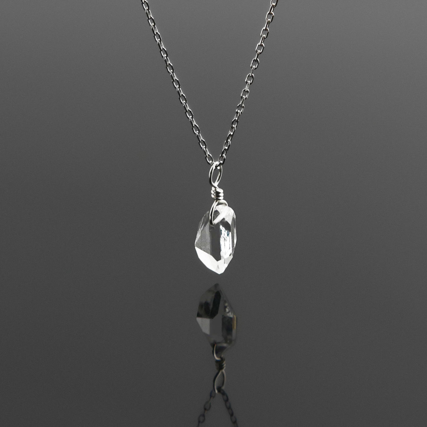 Side view of a dainty, high quality AAA clear raw Herkimer Diamond Quartz wire wrapped sterling silver pendant necklace hanging, with a plain reflective gray background.