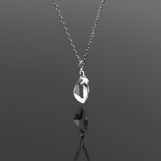 Side view of a dainty, high quality AAA clear raw Herkimer Diamond Quartz wire wrapped sterling silver pendant necklace hanging, with a plain reflective gray background.
