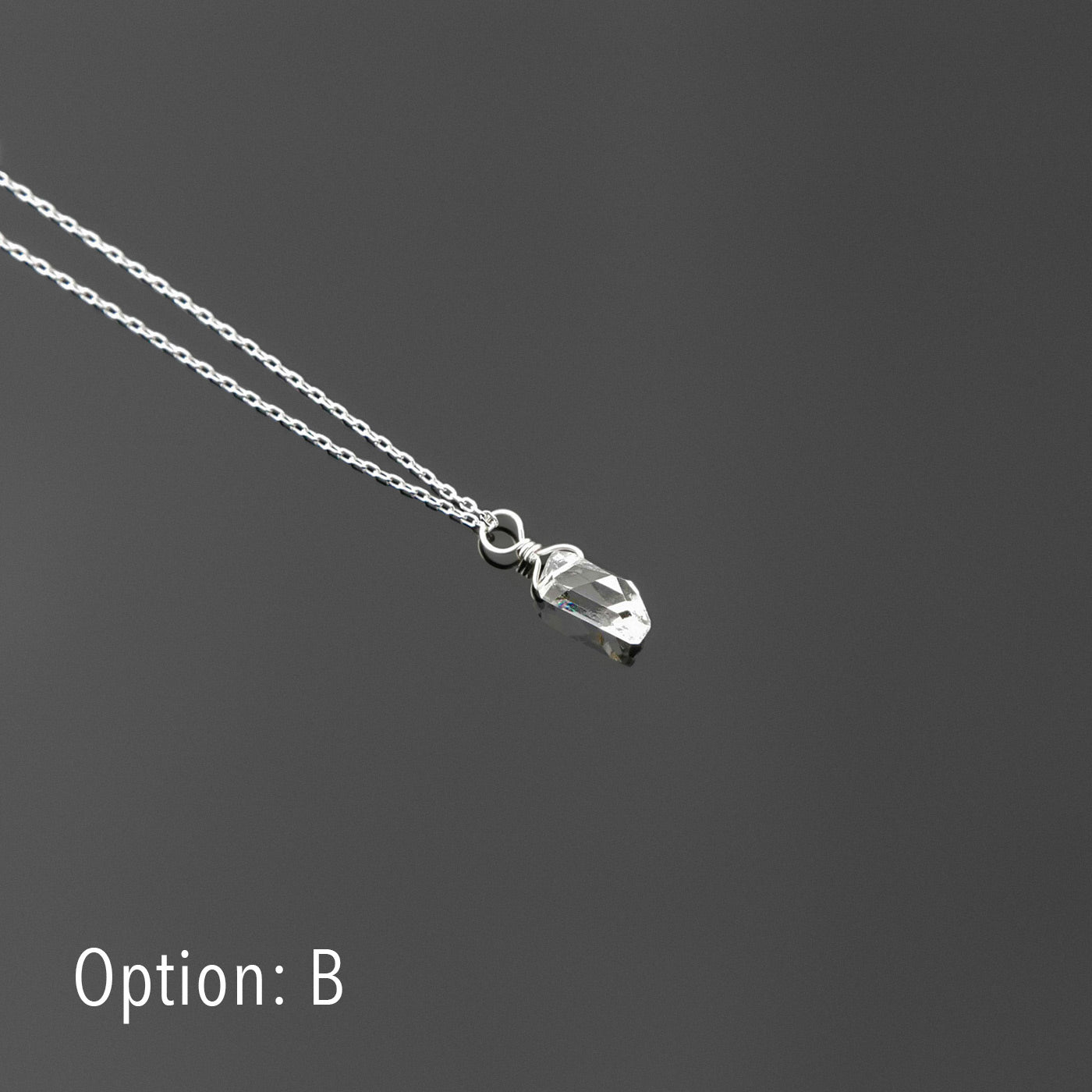3/4 view of a dainty, high quality AAA clear raw Herkimer Diamond Quartz wire wrapped sterling silver pendant necklace, laying flat on a plain reflective gray background.