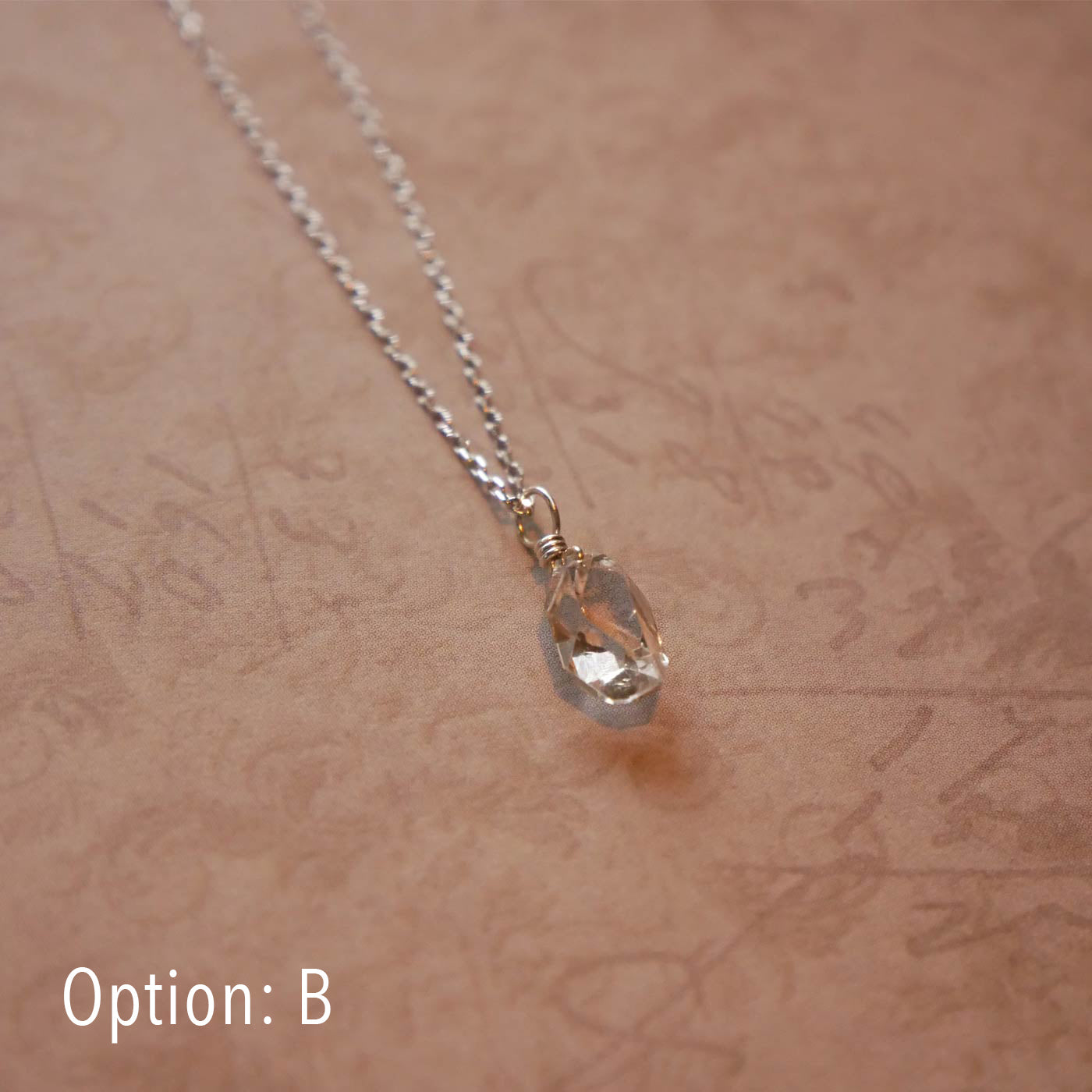 High quality AAA dainty Herkimer Diamond Quartz wire wrapped pendant chain necklace in sterling silver. It is stylized laying flat over vintage patterned pinkish beige paper underneath.