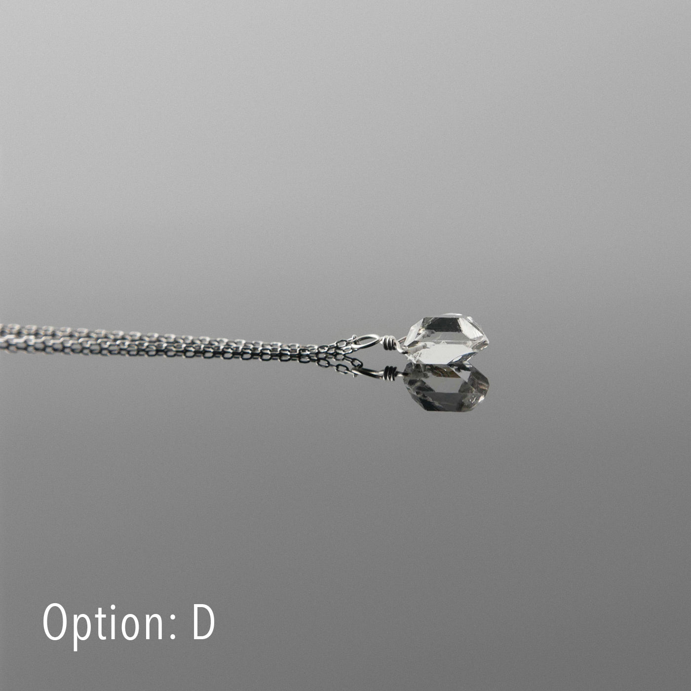 Side view of a dainty, high quality AAA clear raw Herkimer Diamond Quartz wire wrapped sterling silver pendant necklace, laying flat on a plain reflective gray background.
