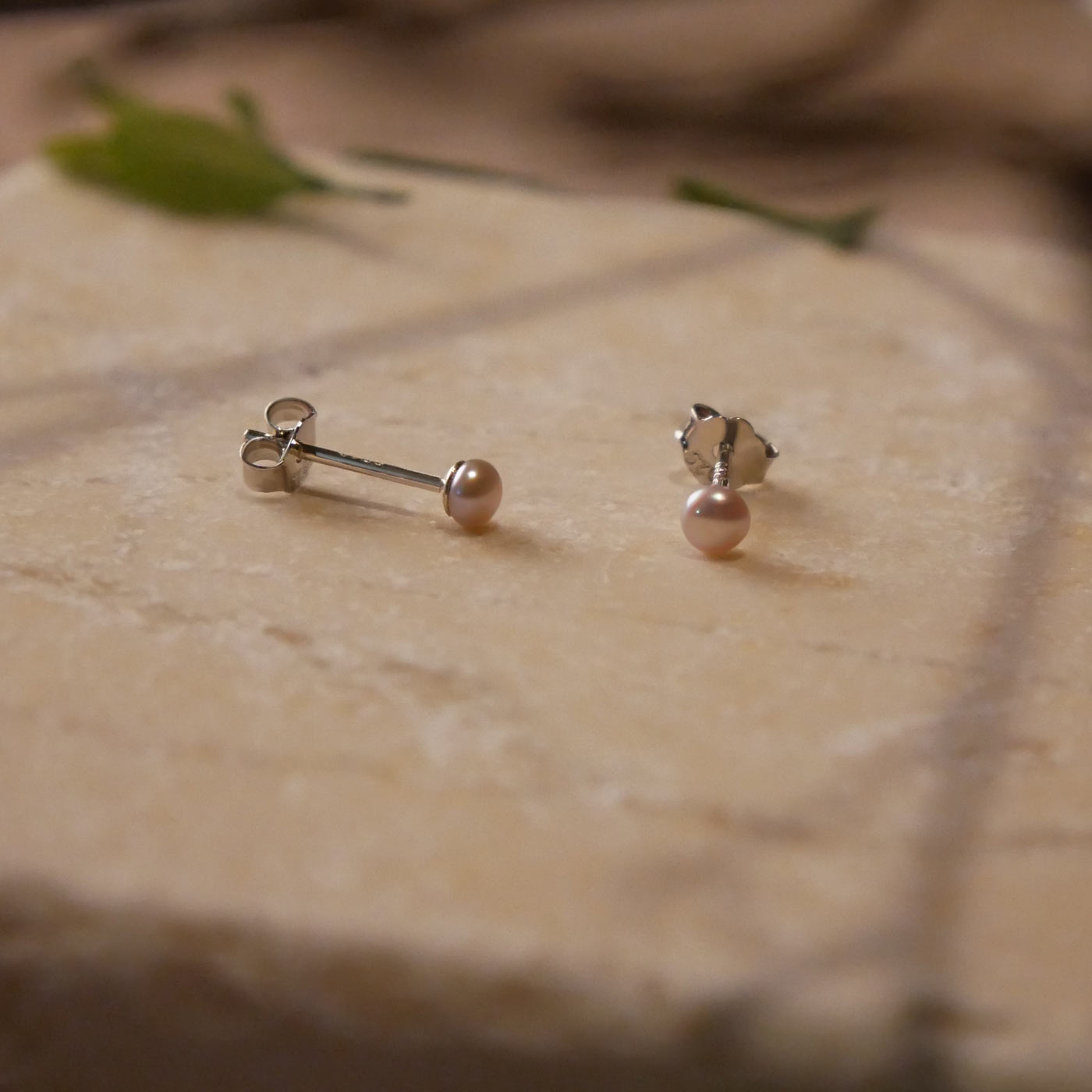 Front and side view of minimalist dainty pair of natural pink pearl sterling silver stud earrings on a stone surface, stylized with  branch shadows and leaves as accents.