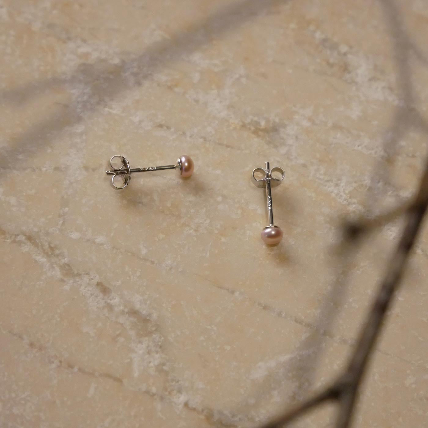 Top view of a minimalist dainty pair of natural pink pearl sterling silver earring studs on a textured stone surface, stylized with branch and branch shadow accents.