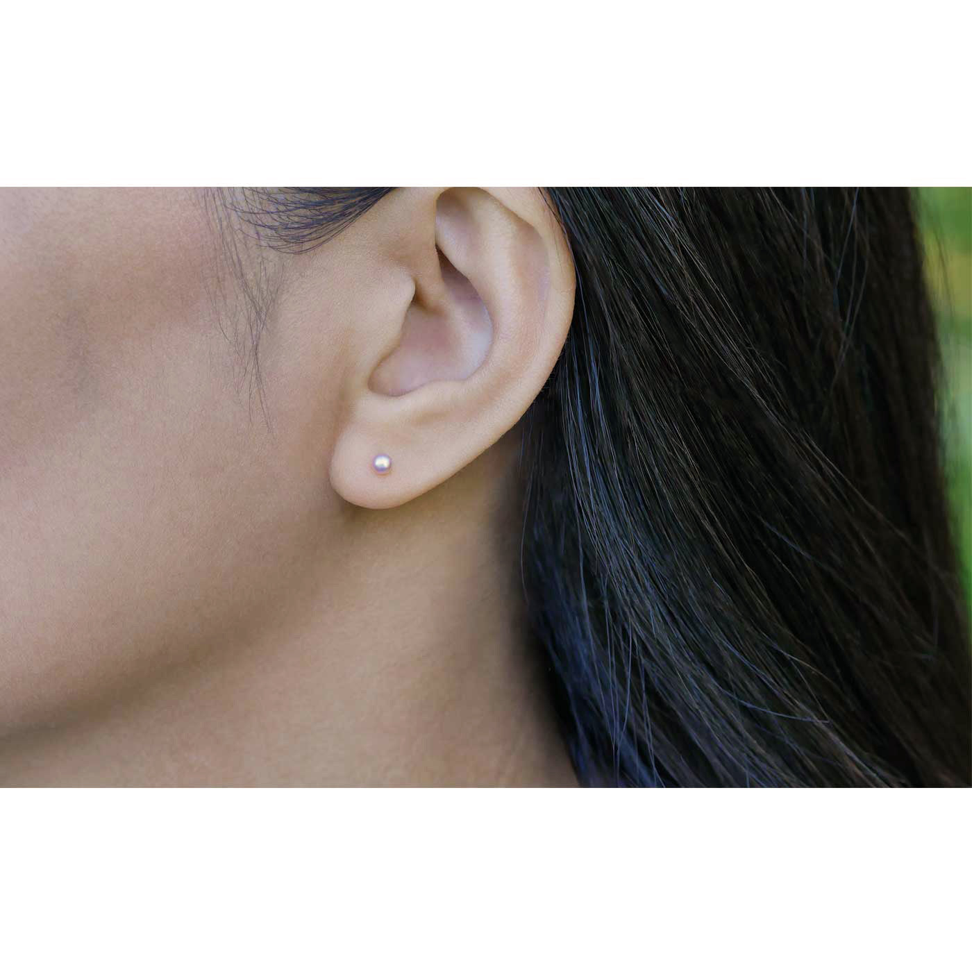 Close-up of dainty minimalist natural pink pearl sterling silver earring worn outside on a person with dark hair, with blurred green plants in the background.