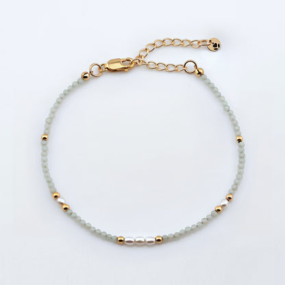 Top view of a dainty smooth round amazonite and accent rice pearls high quality AAA beaded bracelet, with 14K gold filled accent beads, clasp, reverie blossom brand logo tag and extender chain laying flat on a plain off white or light gray background.