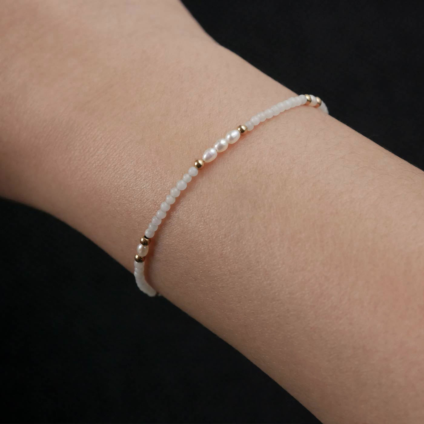 Close-up of a wrist wearing a dainty beaded bracelet with smooth amazonite beads, rice pearls, and 14K gold filled accents. The background is dark black.