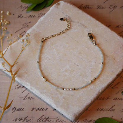 Top view of a dainty beaded bracelet with smooth amazonite beads, rice pearls and 14K gold filled accent beads. It is laying flat on a stone surface stylized with a background of vintage light pink beige paper underneath with old cursive writing, dried beige baby breath flowers and green leaves.
