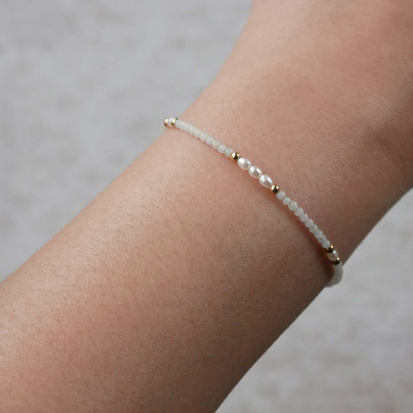 Close-up of a wrist wearing a dainty beaded bracelet with smooth amazonite beads, rice pearls, and 14K gold filled accents. The background is a beige neutral color.