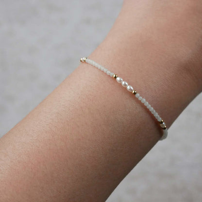 Close-up of a wrist wearing a dainty beaded bracelet with smooth amazonite beads, rice pearls, and 14K gold filled accents. The background is a beige neutral color.