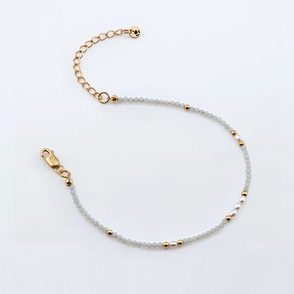 Top view of a dainty smooth round amazonite and accent rice pearls high quality AAA beaded bracelet, with 14K gold filled accent beads, clasp, reverie blossom brand logo tag and extender chain laying flat and open on a plain off white or light gray background.