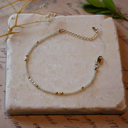 Top 3/4 view of a dainty beaded bracelet with smooth amazonite beads, rice pearls and 14K gold filled accent beads. It is laying flat on a stone surface stylized with a background of vintage light pink beige paper underneath with old cursive writing, dried beige baby breath flowers and green leaves.