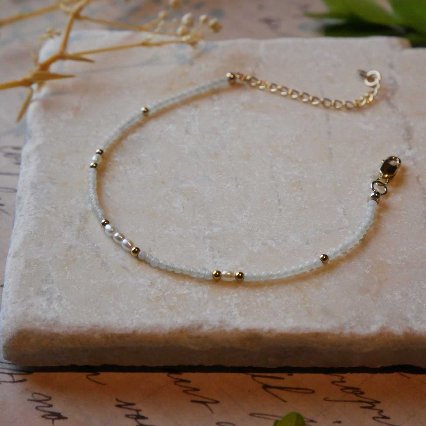 3/4 view of a dainty beaded bracelet with smooth amazonite beads, rice pearls and 14K gold filled accent beads. It is laying flat on a stone surface stylized with a background of vintage light pink beige paper underneath with old cursive writing, dried beige baby breath flowers and green leaves.